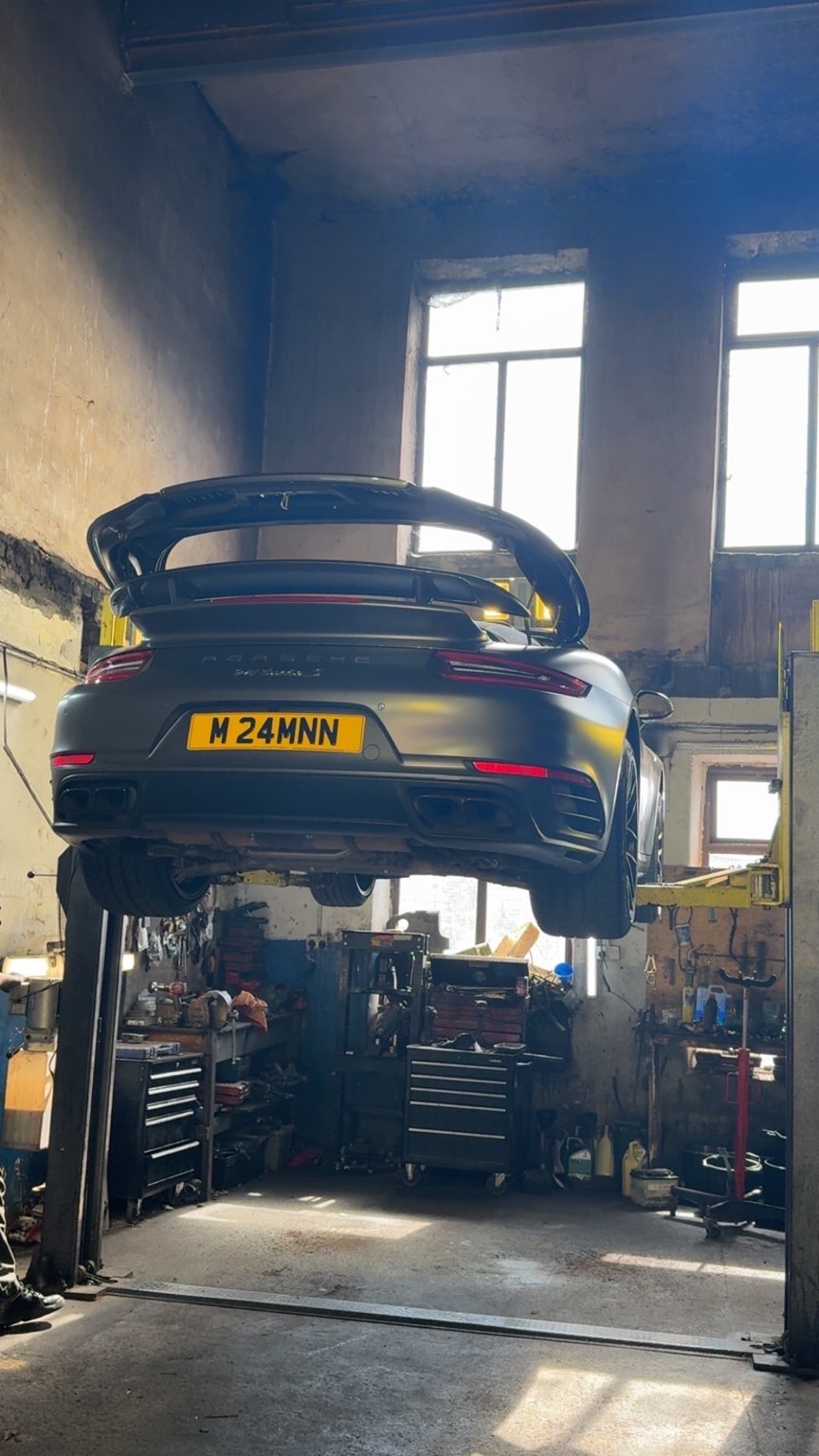 A Porsche 911 Turbo S raised on the two-post lift inside the stone workshop, with light coming through the tall mill windows
