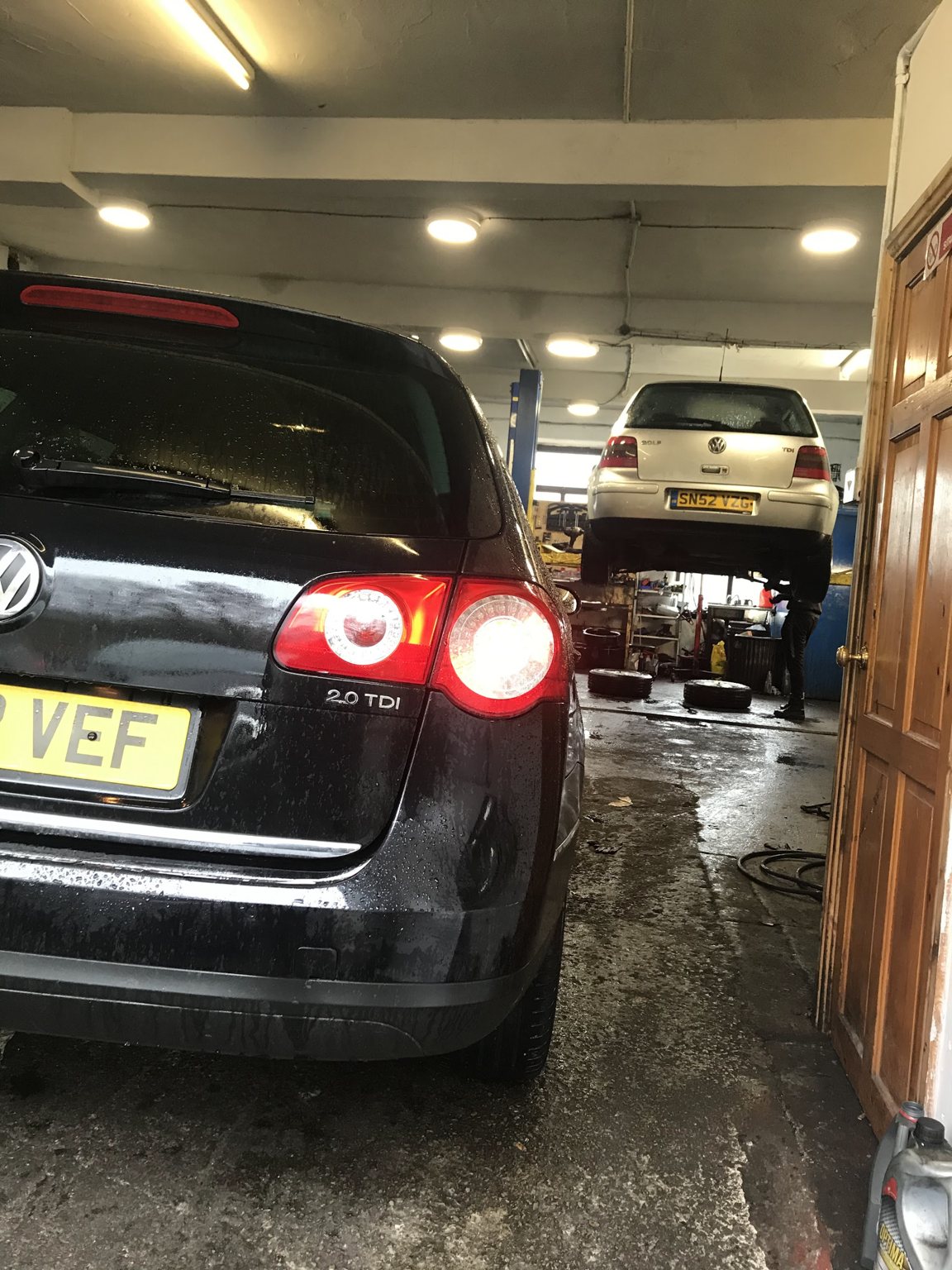 Inside the workshop: a Volkswagen estate waiting at the door while a Golf is up on the lift with a mechanic working underneath